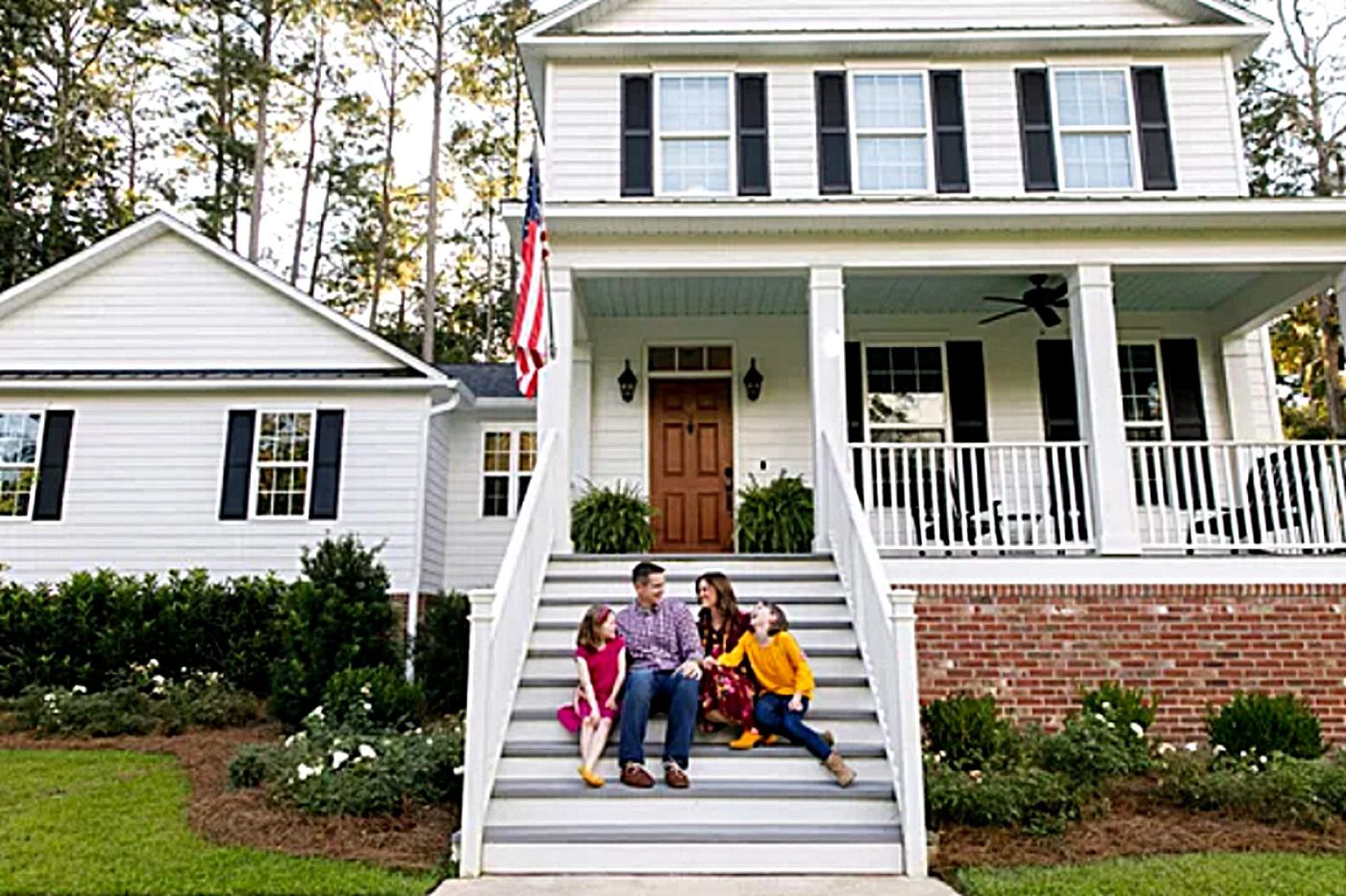 A man and a woman sit on the front steps of a white two-story house with their two children on a bright afternoon
