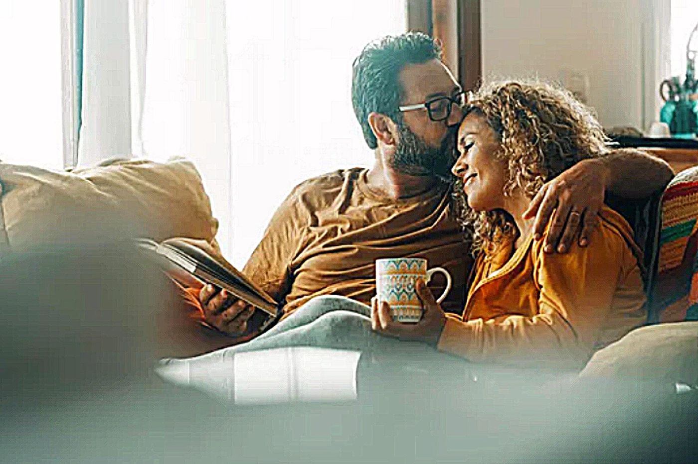 A man and a woman sitting together on a couch at home in warm morning light, one holding a coffee mug, talking over an open notebook