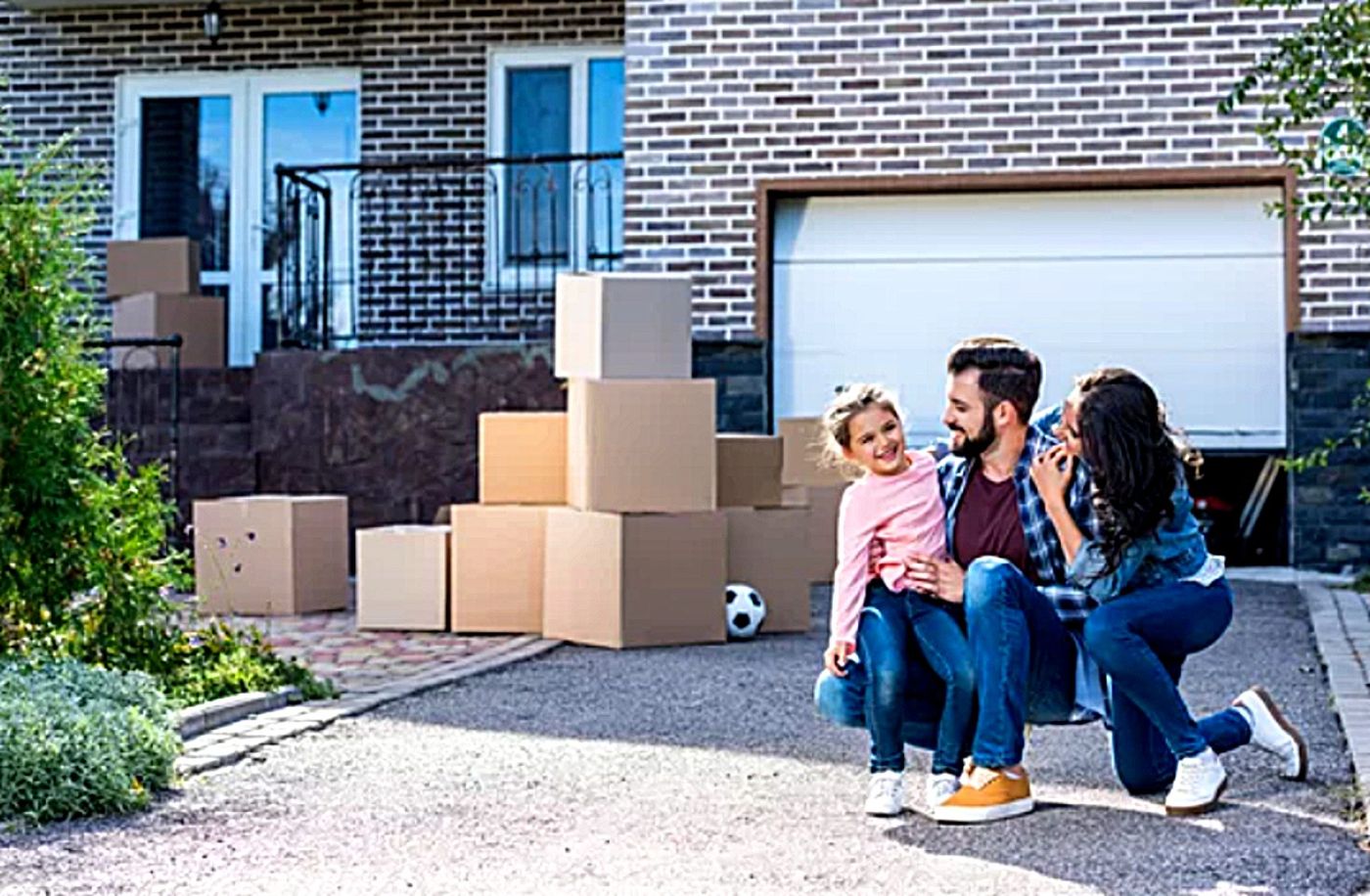 A man and a woman kneeling in a driveway with their young daughter, moving boxes stacked behind them on the day they take possession of a first home