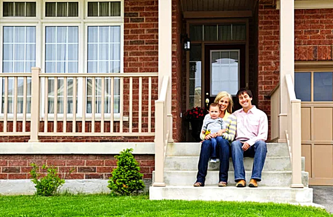 A man and a woman sit on the front steps of a brick house with their young child on a sunny day